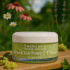 Een persoon die het Éminence Organic Skin Care Citrus & Kale Potent C+E Masque potje vasthoudt, in een natuurlijke buitenomgeving met groen op de achtergrond.